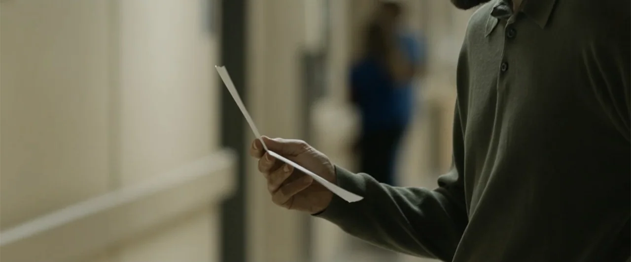 Unsung Hero (2024) movie still: medium — A man stands in a sterile institutional corridor with light walls and blurred background elements. He holds…
