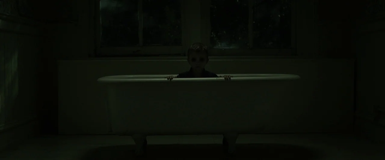 Before I Wake (2016) movie still: medium — A dark bathroom is dominated by a white claw-foot tub against a window. A young child with solid black…