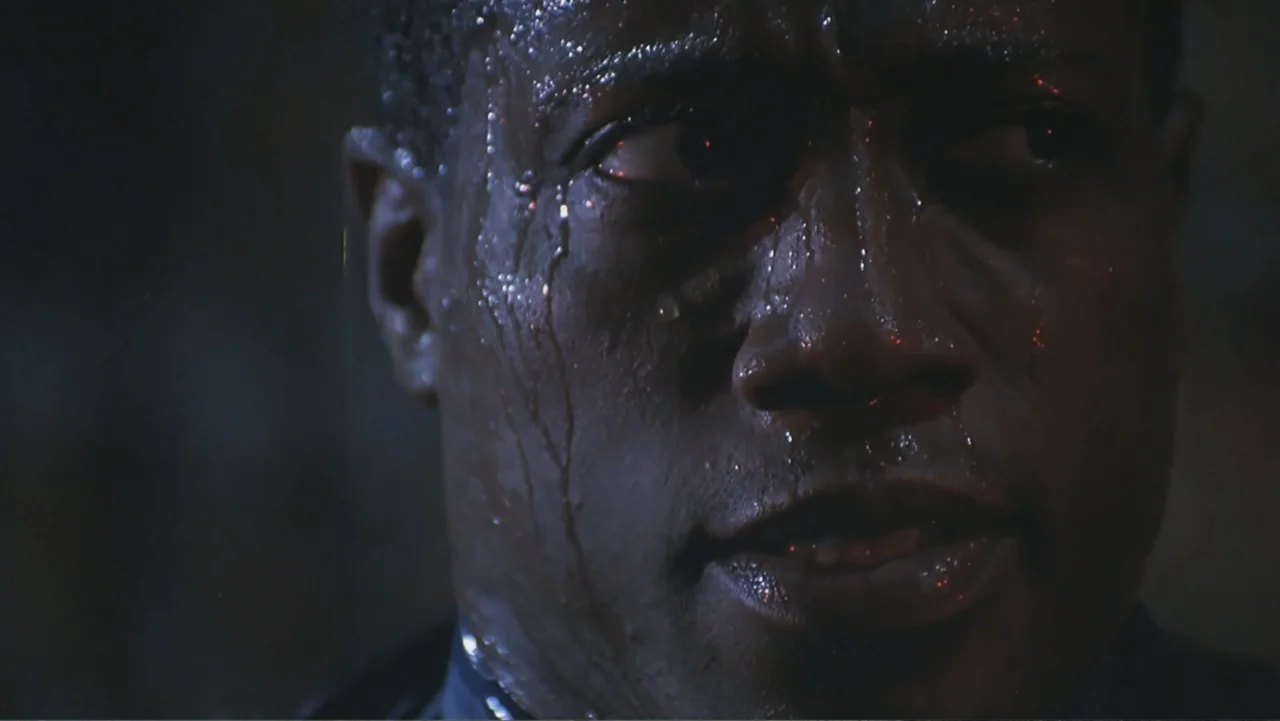 The Art of War (2000) movie still: closeup — A dark, indistinct interior setting with minimal background details visible in shadow. A man with beads…
