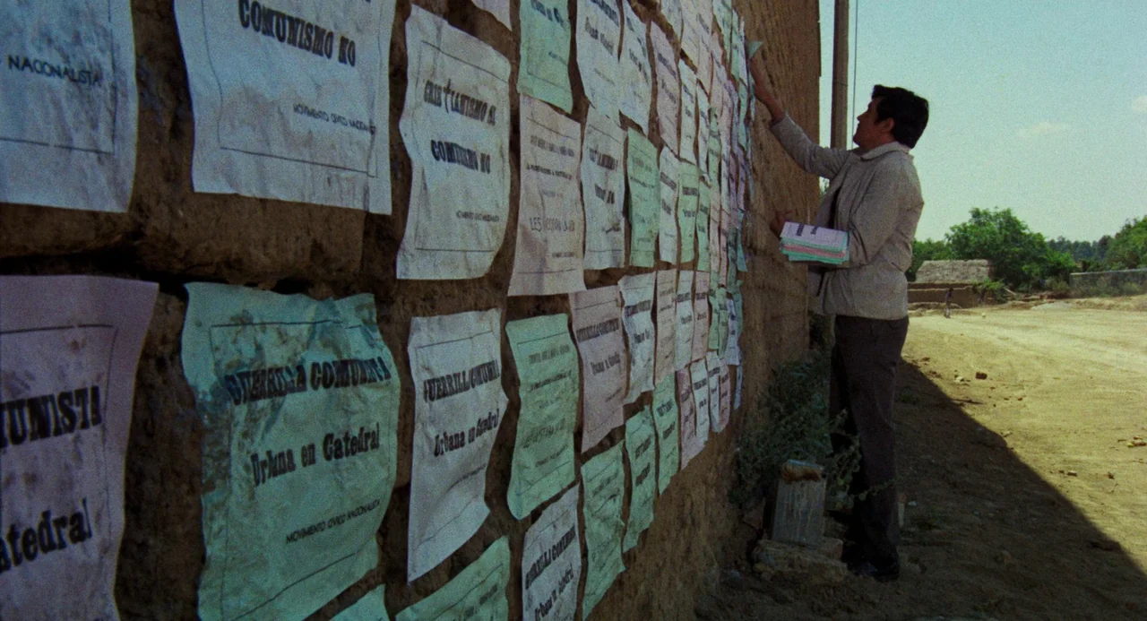 Canoa: A Shameful Memory (1976) movie still: fullbody — A man tapes various colorful flyers to a rough, rustic adobe brick wall. The wall stands…