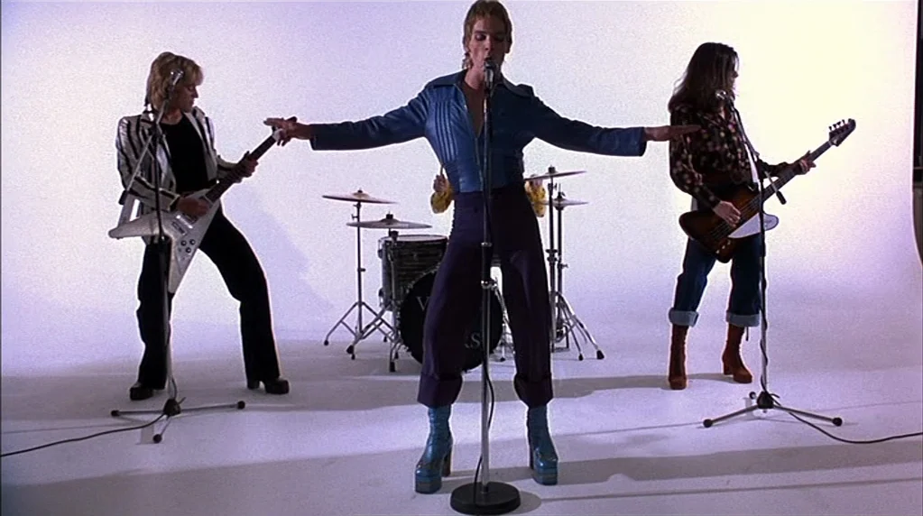 Velvet Goldmine (1998) movie still: fullbody — A minimalist white studio background surrounds a musical group on a sound stage. The band members…