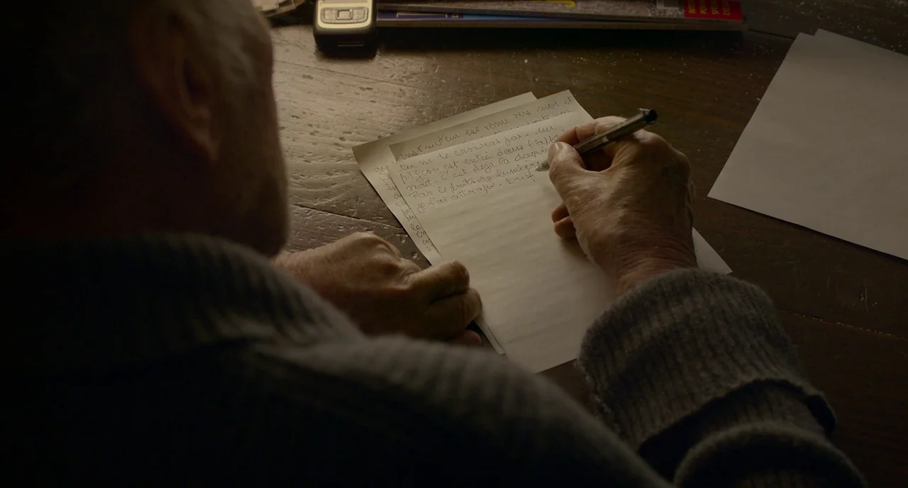 Amour (2012) movie still: closeup — A wooden table surface cluttered with a mobile phone and magazines lies under lighting. An elderly person writes…
