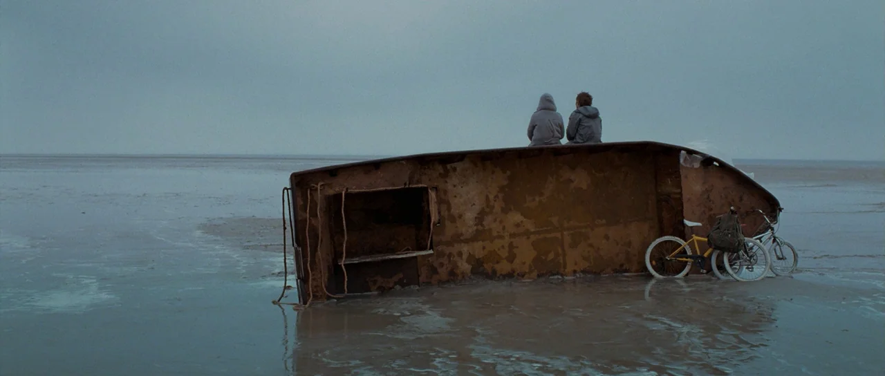 Womb (2010) movie still: wide — A vast, flat expanse of mud stretches to the horizon under a grey sky. Two young males sit on the side of a large…