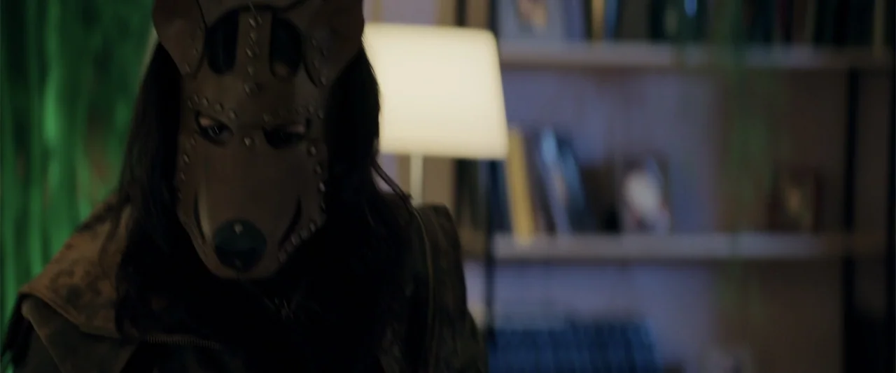 Human Animals (2020) movie still: closeup — A dimly lit room contains a blurred bookshelf in the background under soft lamp light. A person wearing…
