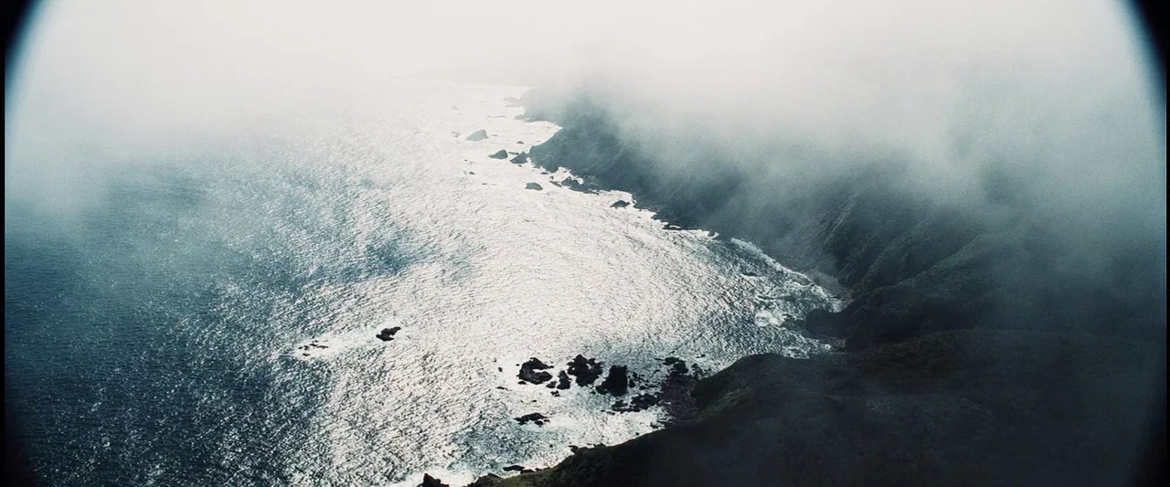 The Island (2005) movie still: establishing — A rugged coastline stretches along a dark sea beneath dense, low-hanging ocean mist. The terrain…