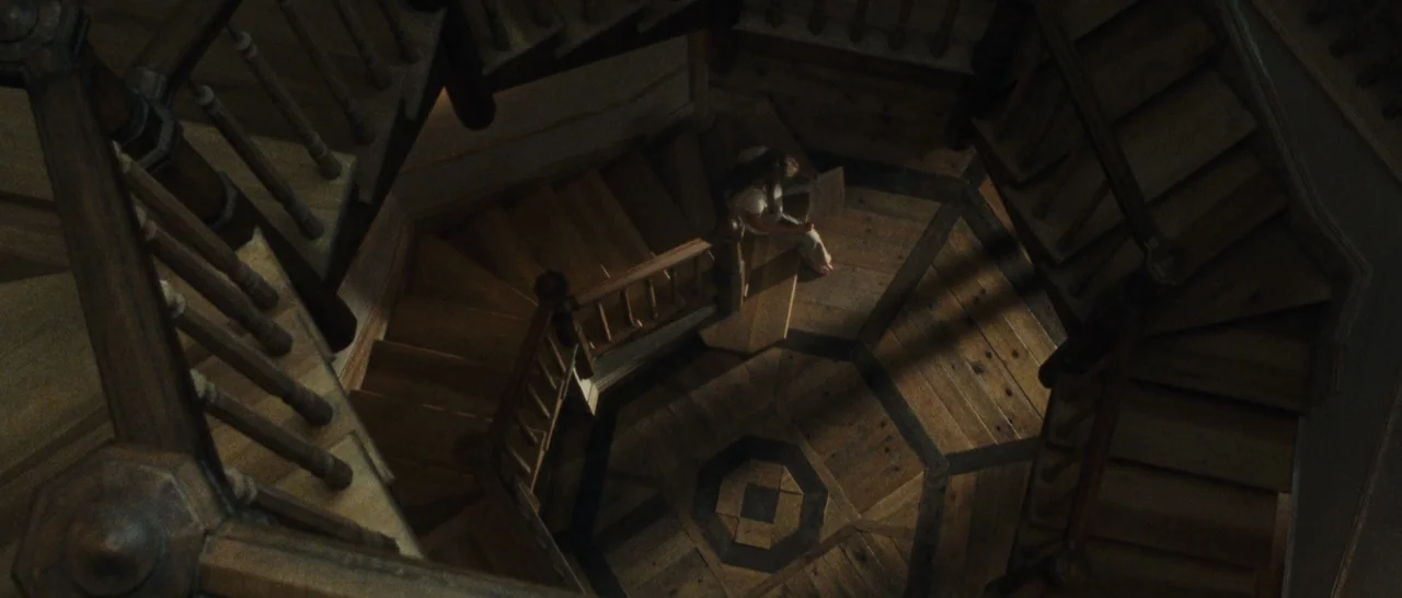 mother! (2017) movie still: wide — A dark wooden octagonal staircase winds down into a central landing area. A woman sits on the steps reading a…