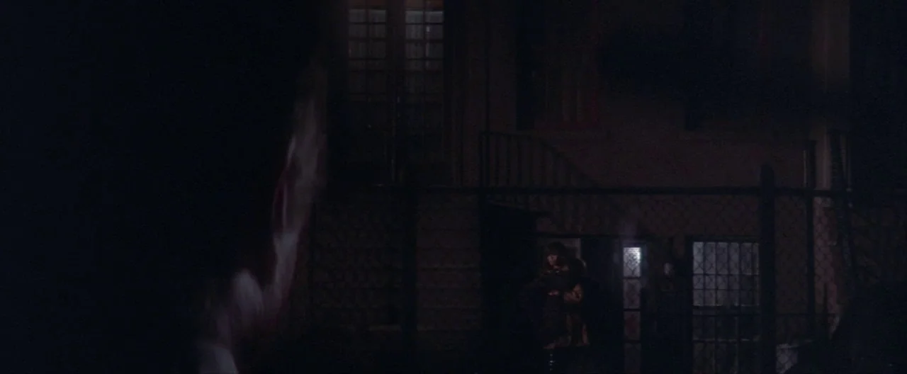 Klute (1971) movie still: wide — A dark outdoor urban space features a fenced perimeter and a dimly lit doorway. A woman stands in the entrance…