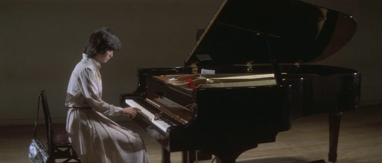 Take Me Away! (1978) movie still: medium — A dark, spare practice room contains only a black grand piano and a wooden chair. A young woman in a…