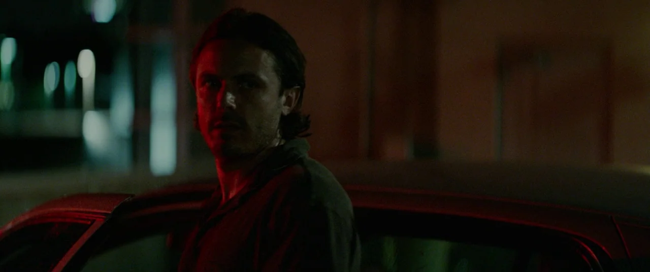 Triple 9 (2016) movie still: medium — A man stands beside an open car door in a dimly lit, dark parking lot. The man looks toward the left side of…
