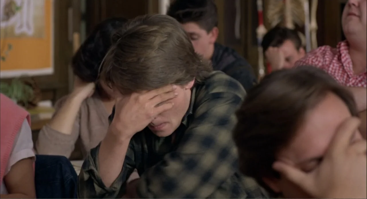 Teen Wolf (1985) movie still: medium — A high school classroom contains several students sitting at desks in rows. The students hold their hands…
