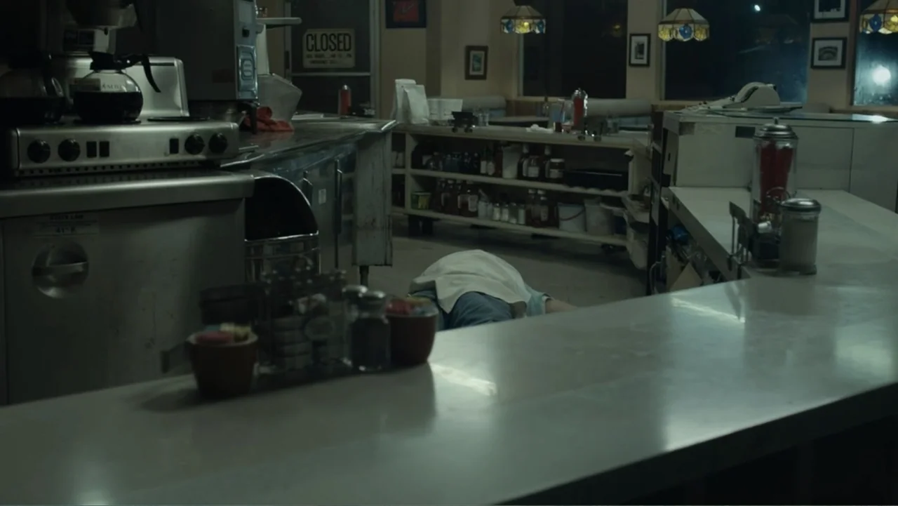 The Cleaner (2021) movie still: wide — A dark, empty diner interior features a long service counter and stainless steel appliances. A person lies…