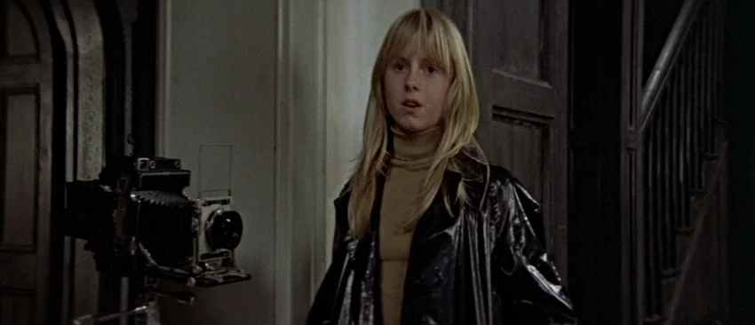 Images (1972) movie still: medium — A dimly lit residential foyer features dark wooden doors and a staircase. A young girl with blonde hair stands…