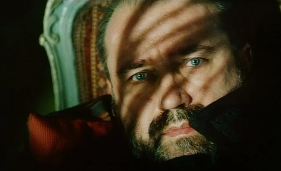 Simon, The Magician (1999) movie still: closeup — A man sits in a patterned fabric chair while illuminated by striped light. The sharp contrast…