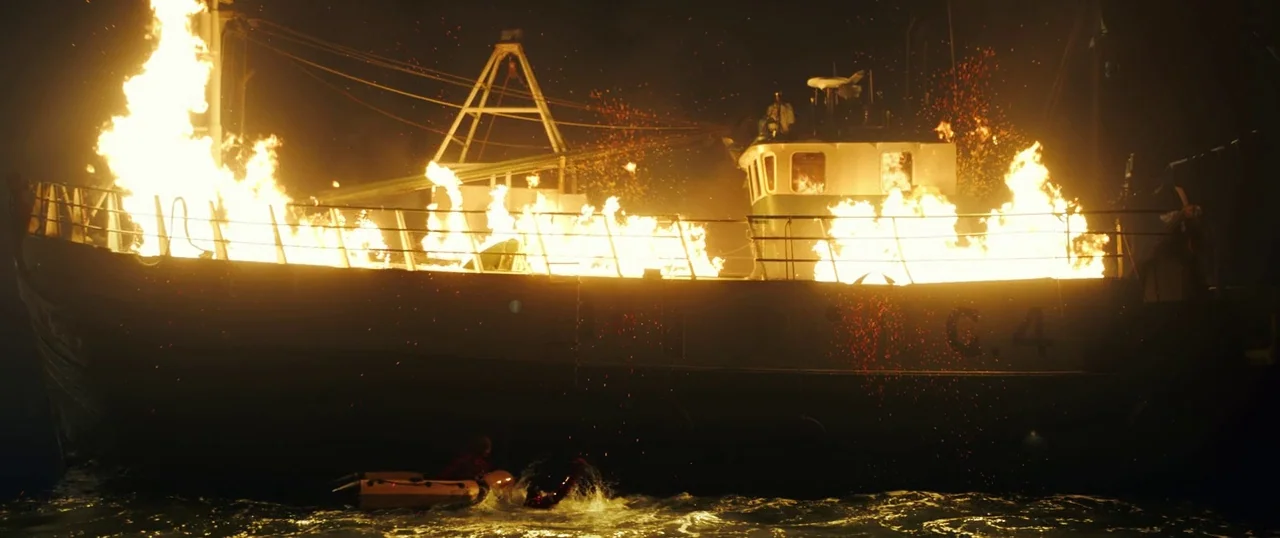 Sea Fever (2019) movie still: wide — A fishing trawler burns intensely on dark ocean water at night. Sparks and embers rise from the deck into the…