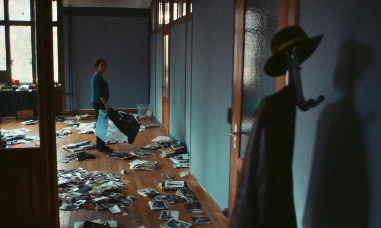 Little Girl Blue (2023) movie still: wide — A long hallway with blue walls and parquet wood floors covered in scattered photographs. A woman stands…
