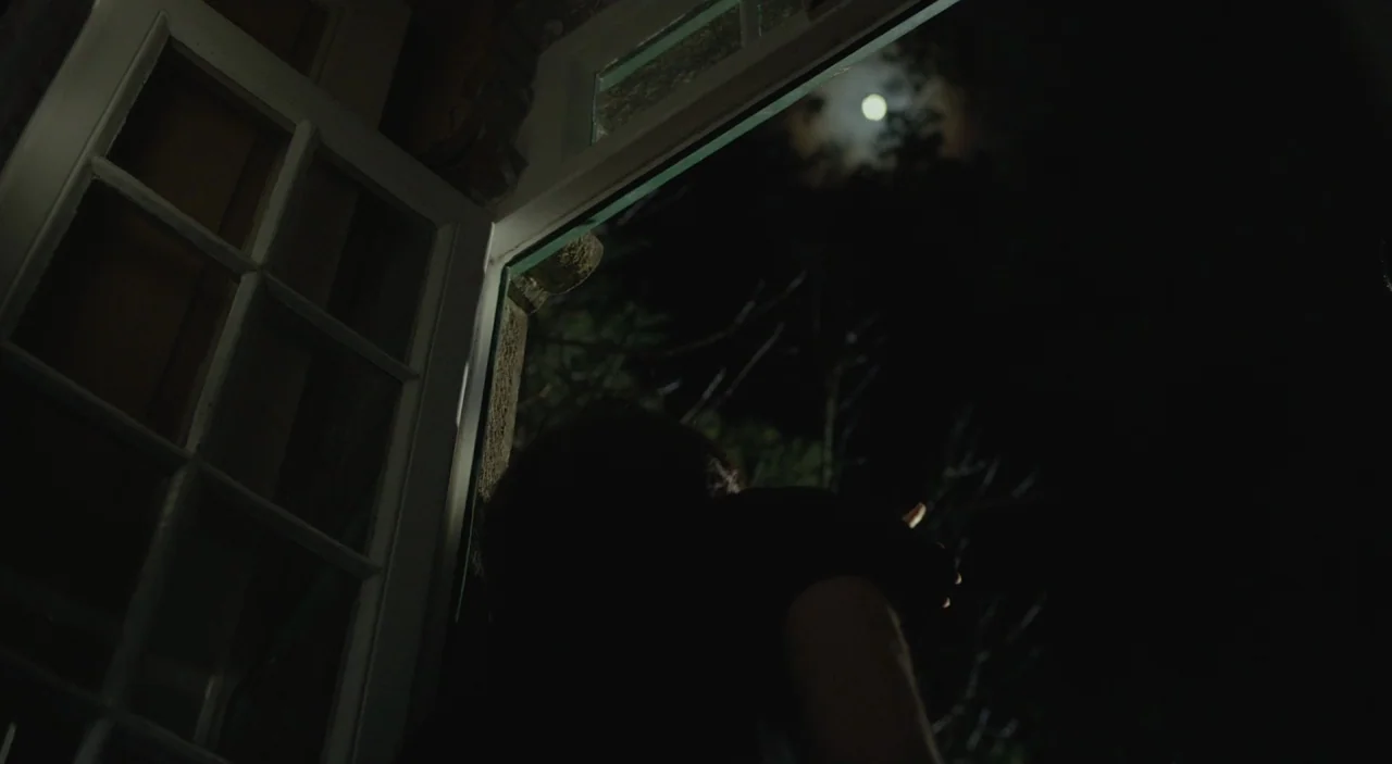Cosmos (2015) movie still: medium — A dark bedroom with an open sash window looking out onto a night sky. A person stands facing the window,…