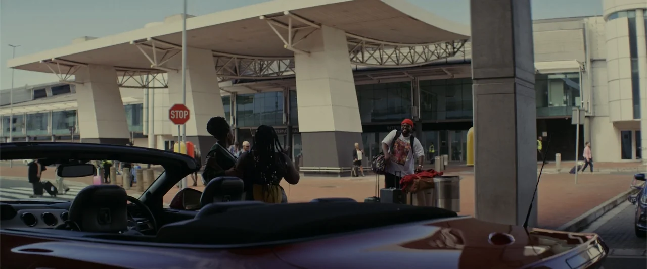 A Scam Called Love (2025) movie still: medium — Modern airport terminal drop-off area features large white architectural supports and glass facades.…