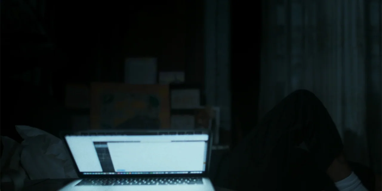 Nokturno (2024) movie still: medium — A dark bedroom interior is lit by the blue glow of a computer screen. A person with legs pulled up sits…