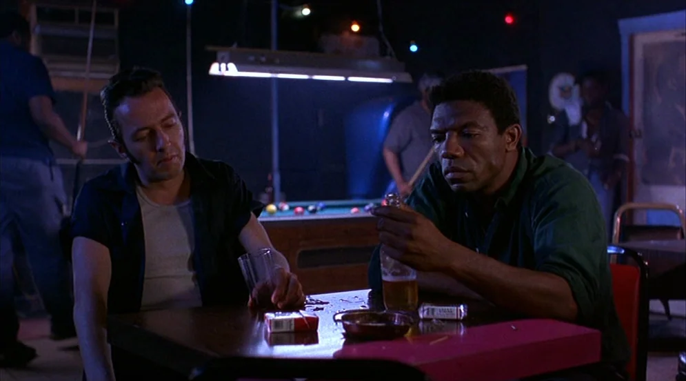 Mystery Train (1989) movie still: medium — A dark, low-lit billiard hall with people standing near pool tables in the background. Two men sit at a…