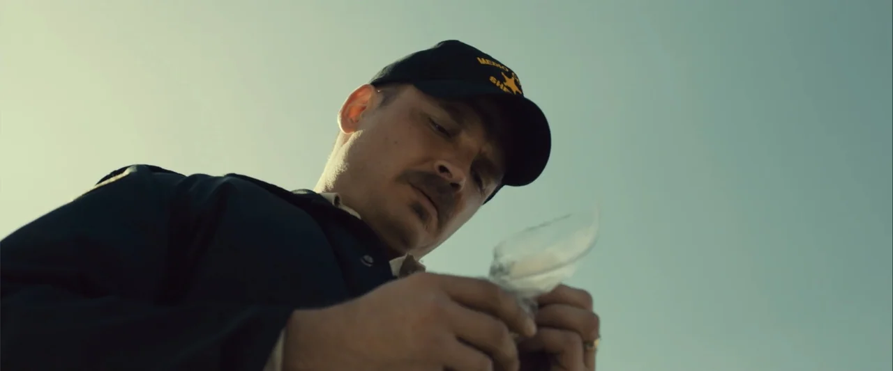 Frontera (2014) movie still: closeup — An outdoor desert location with a clear bright sky overhead. A man in a law enforcement uniform inspects a…