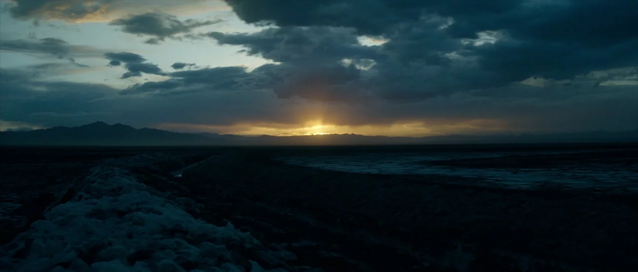 The Neon Demon (2016) movie still: establishing — A vast salt flat extends toward a distant mountain range under a stormy sky. The sun sets low on…