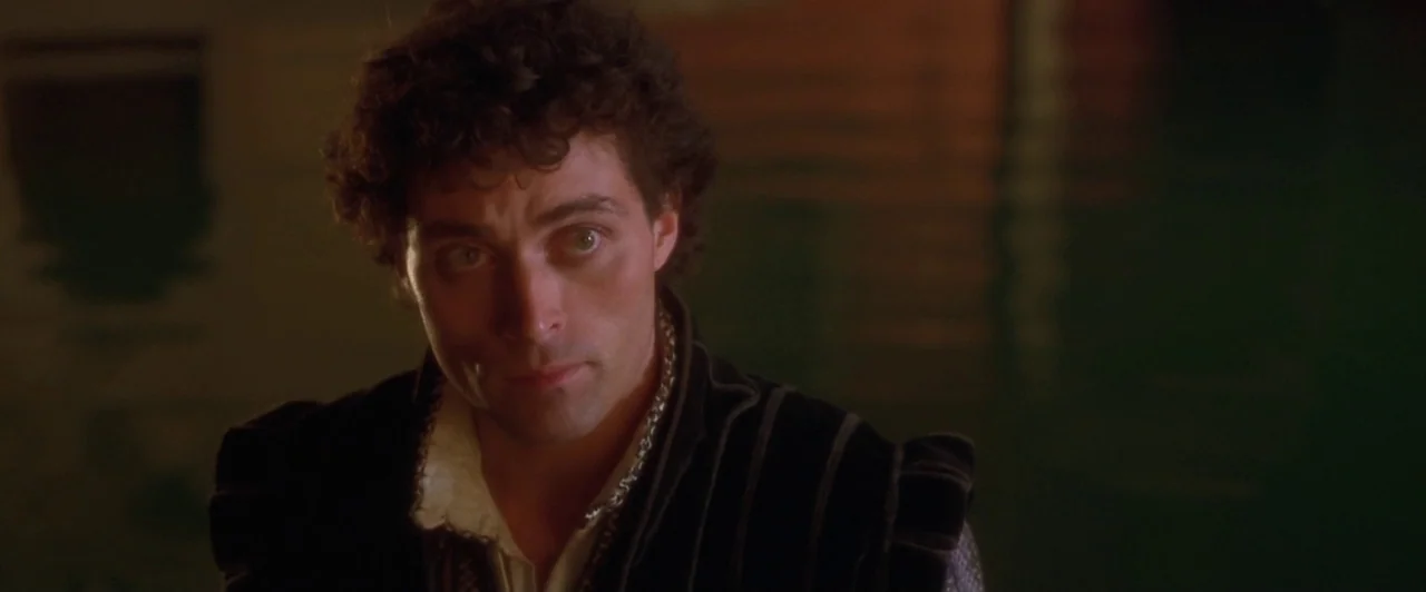 Dangerous Beauty (1998) movie still: closeup — A man stands before dark water reflecting amber light at night. He stares ahead while wearing a dark…