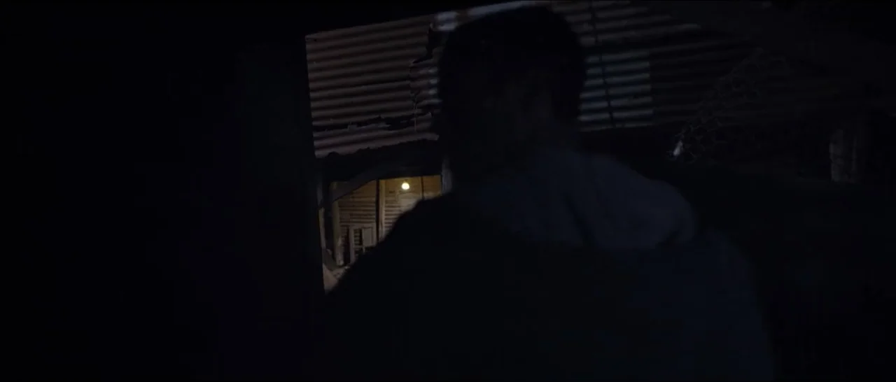 Gunman (2025) movie still: medium — An interior space with a corrugated metal wall and a dimly lit background area. A person seen from behind stands…