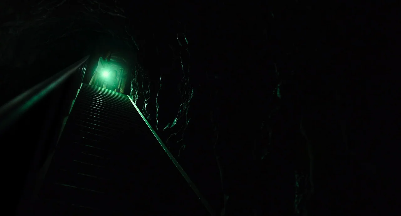The Mimic (2017) movie still: wide — A long flight of wooden stairs leads into darkness within a rock cavern. A bright green light shines from the…