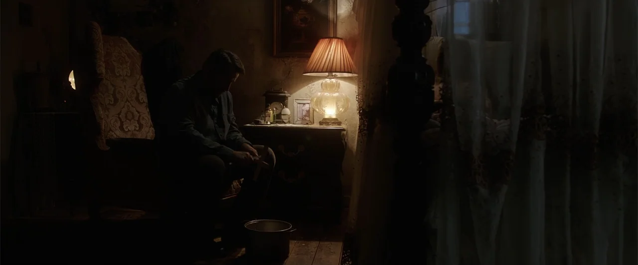Annabelle: Creation (2017) movie still: medium — A dim bedroom contains a wooden armchair and a side table with a lamp. A man sits in the chair and…