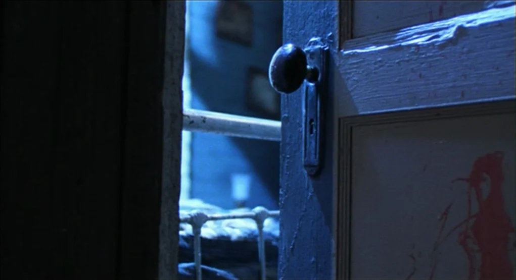 Evil Dead II (1987) movie still: closeup — A partially open wooden door shows a glimpse of a shadowed, dimly lit room. A splash of bright red blood…