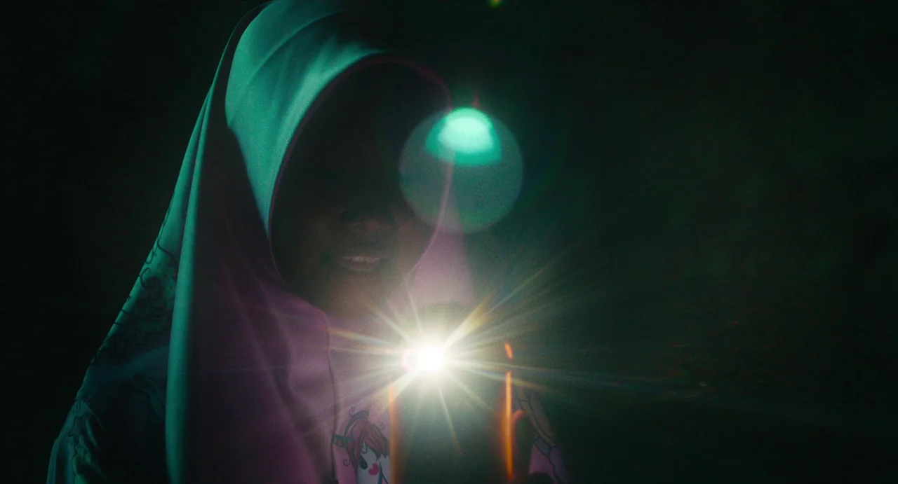 Tiger Stripes (2023) movie still: closeup — A person wearing a pink headscarf is positioned in a dark environment. They hold a lit flashlight…