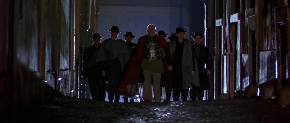 Titus (1999) movie still: fullbody — A group of men in hats and coats walks down a dark, narrow cobblestone passage. The central figure wears a…