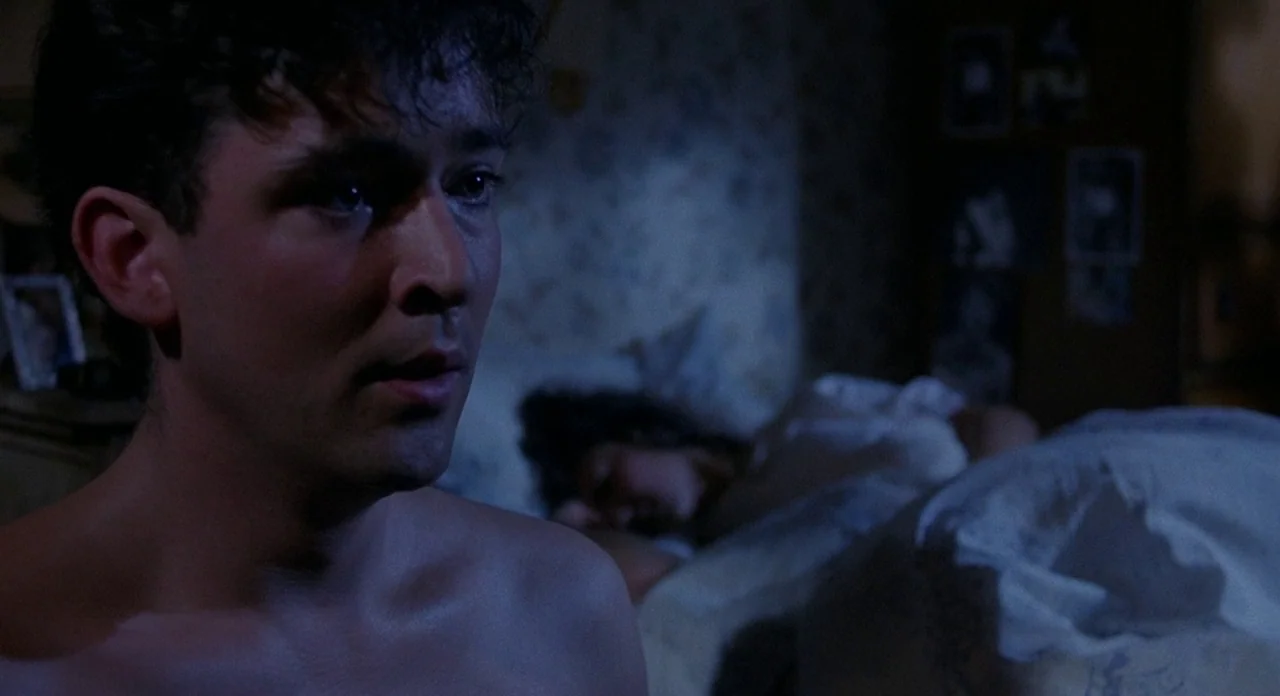 Hellraiser (1987) movie still: closeup — A dimly lit room with wallpapered walls and pictures scattered in the background. A man sits awake in bed…
