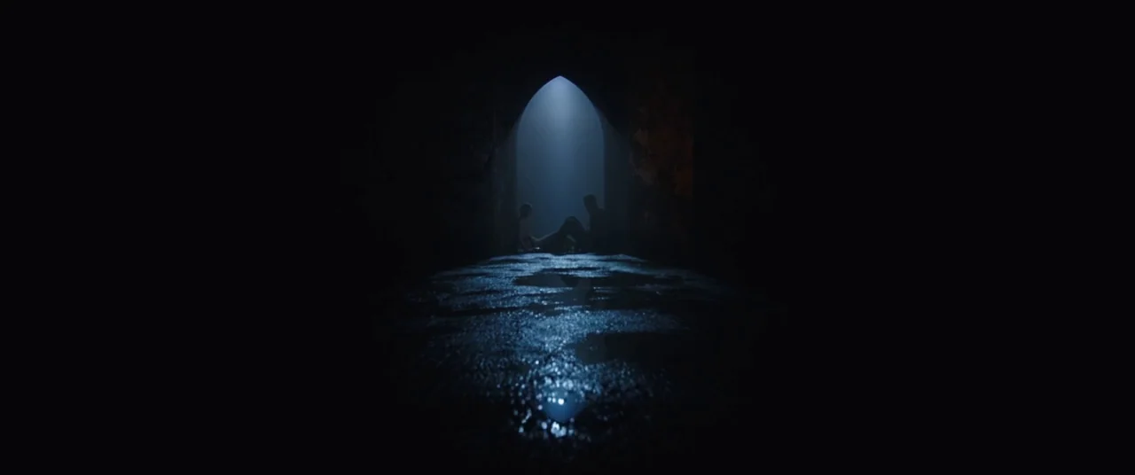 The Nun (2018) movie still: wide — A dark, wet tunnel features a central arched exit glowing with blue light. Two silhouettes sit on the floor of…