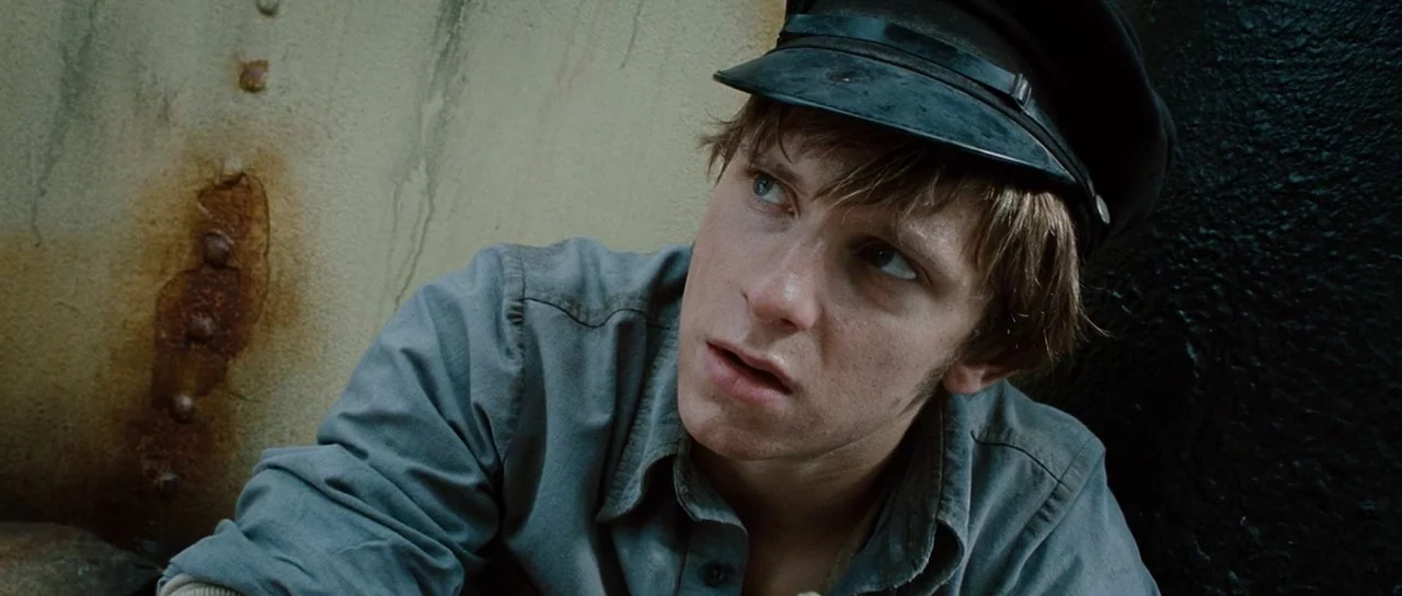 King Kong (2005) movie still: medium — A rusted metal wall with visible oxidation marks forms the background. A young man wearing a service cap…