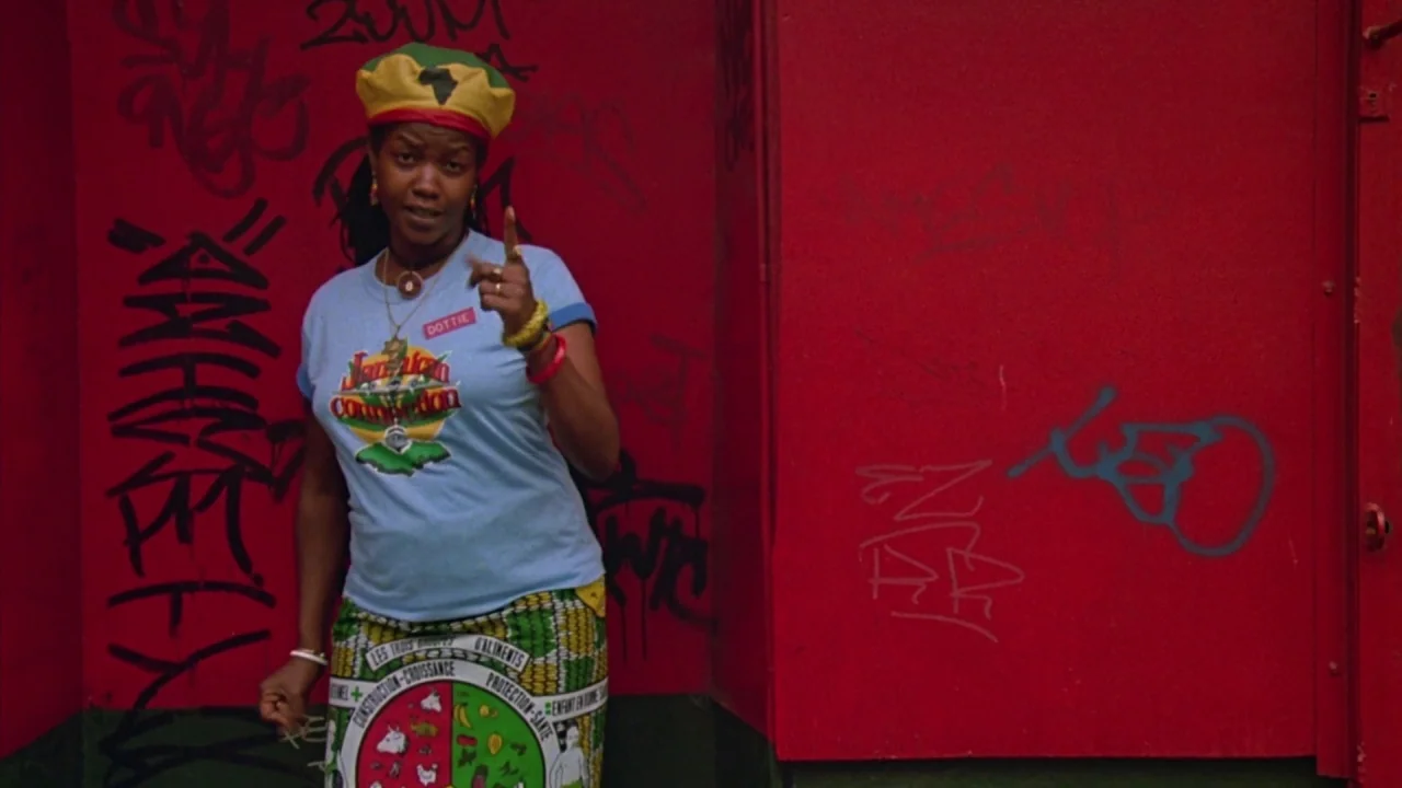 Something Wild (1986) movie still: medium — A woman stands in front of a bright red wall covered in black graffiti markings. She wears a colorful…