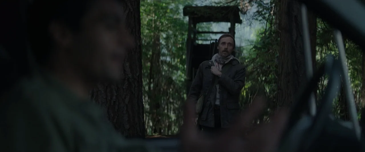 Wolf Man (2025) movie still: medium — A dense forest environment features tall trees and a wooden elevated structure nearby. A man wearing a jacket…
