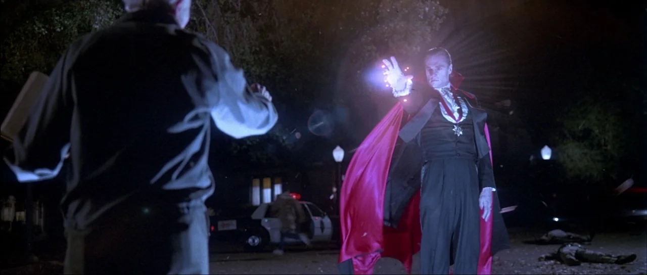 The Monster Squad (1987) movie still: wide — A dark residential street at night features a vampire standing in front of a police car. The vampire…