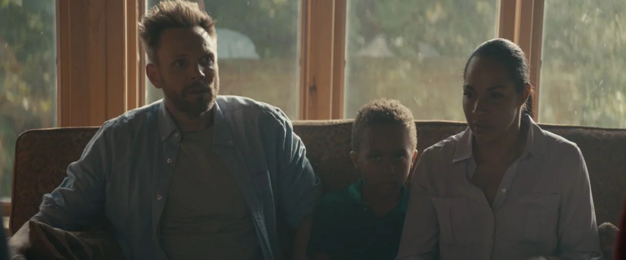 Becky (2020) movie still: medium — A family sits on a patterned couch in front of a multi-pane window. The group watches something to their left…