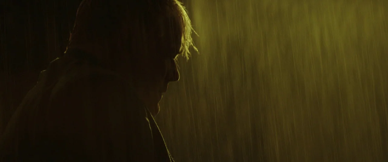 God’s Pocket (2014) movie still: closeup — A man stands in the rain at night under a saturated yellow light. He looks downward while water streaks…
