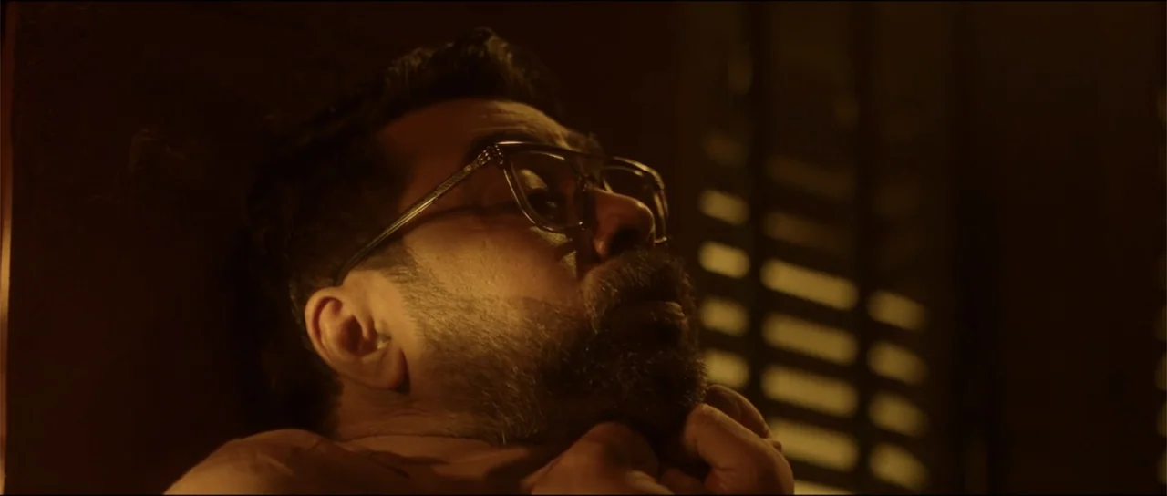 Garudan (2024) movie still: closeup — A dark interior room with slatted blinds visible in the background shadow. A man with a beard and glasses…