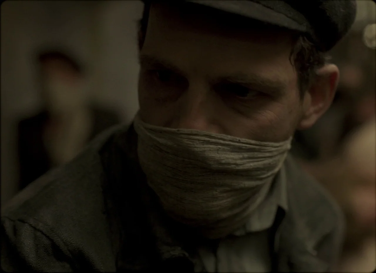Son of Saul (2015) movie still: closeup — A dimly lit room with muted wall tones suggesting an older, austere interior space. A man with a covered…