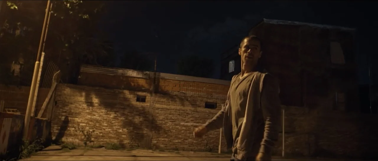 Gunman (2025) movie still: medium — An outdoor urban alleyway at night features a weathered brick wall and sparse foliage. A man wearing a grey…
