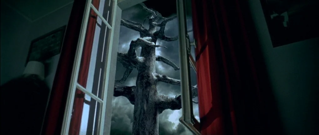 Mortal Transfer (2001) movie still: establishing — A dark, bare tree stands outside a window frame against a cloudy night sky. Red fabric curtains…