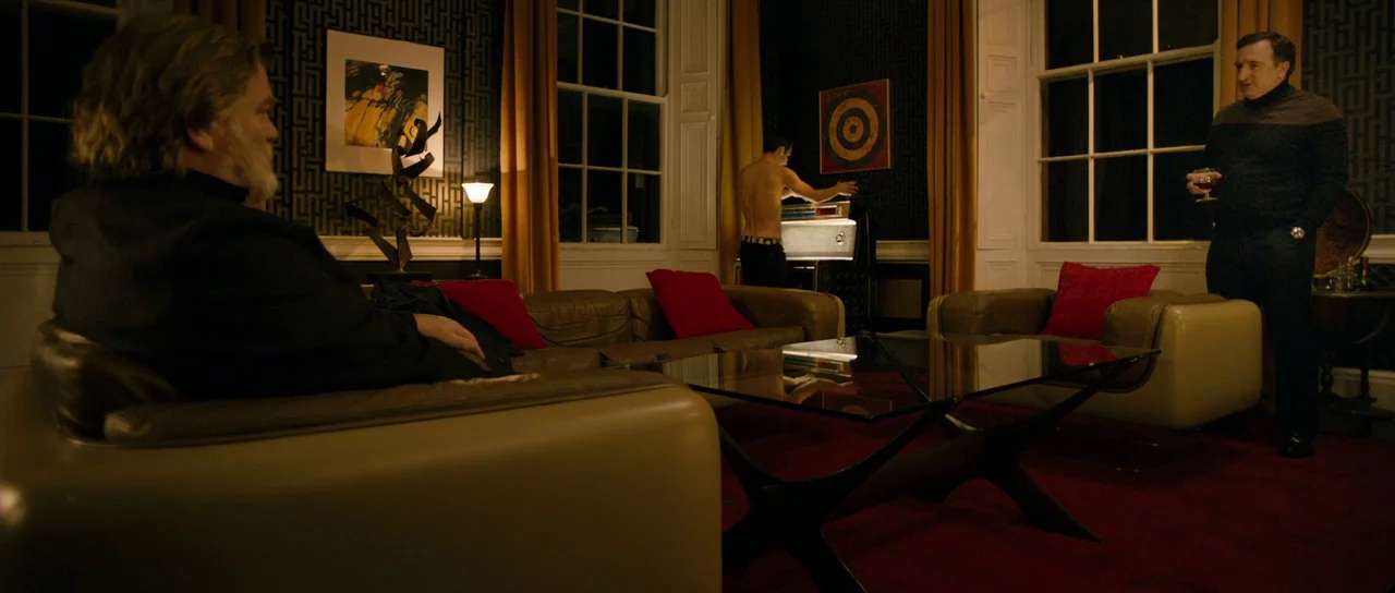 Calvary (2014) movie still: wide — A dark, affluent living room features a patterned wallpaper and mid-century modern furniture pieces. One man sits…