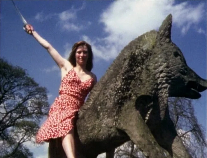 Glitterbug (1994) movie still: fullbody — A woman sits atop a large stone wolf sculpture in an outdoor area. She holds a thin metal sword upward…