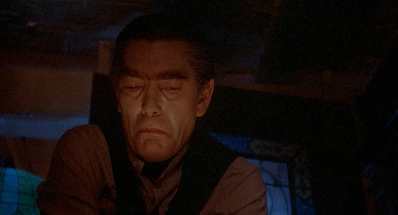 Inferno (1980) movie still: medium — A dimly lit, shadowed room features hints of dark wood and stained glass. A middle-aged man stares downward…
