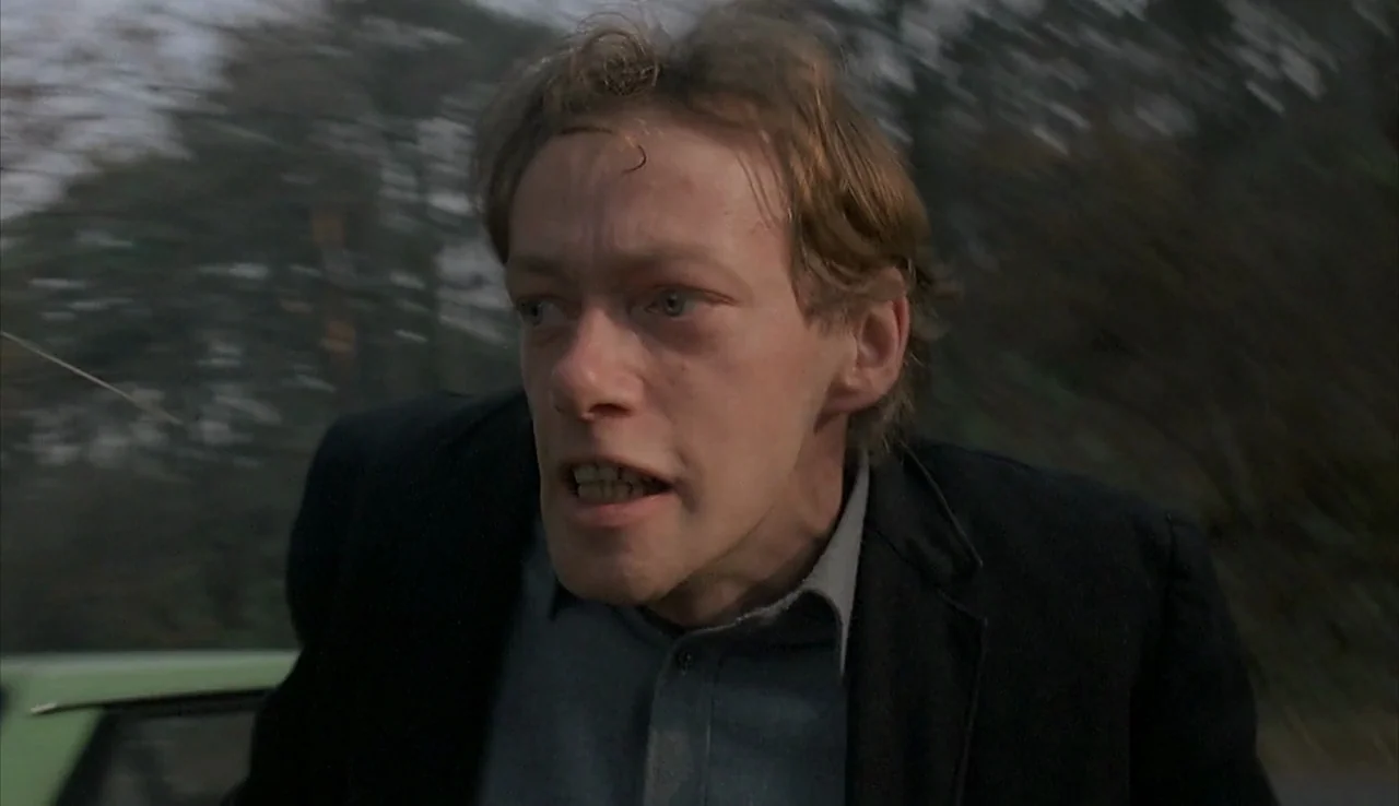 Angst (1983) movie still: closeup — A man stands outdoors near a blurred background of trees and a vehicle. He stares intensely while his hair blows…