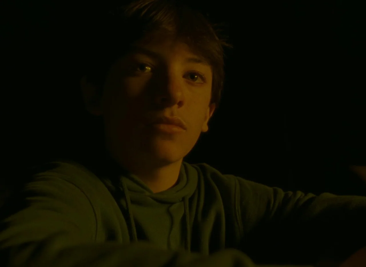 Falcon Lake (2022) movie still: closeup — A darkened interior space with no visible background details. A teenage male looks forward under warm,…