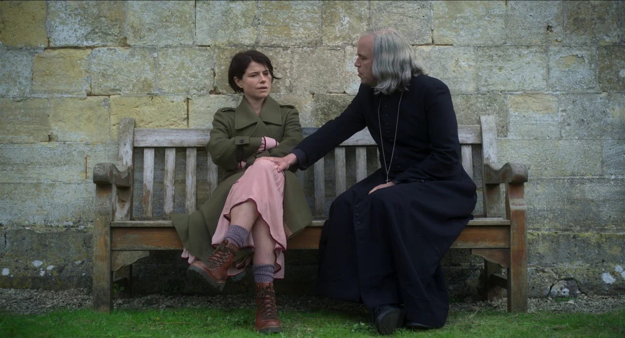 Men (2022) movie still: medium — Two individuals sit together on a wooden bench against a weathered sandstone block wall. A woman looks toward a man…